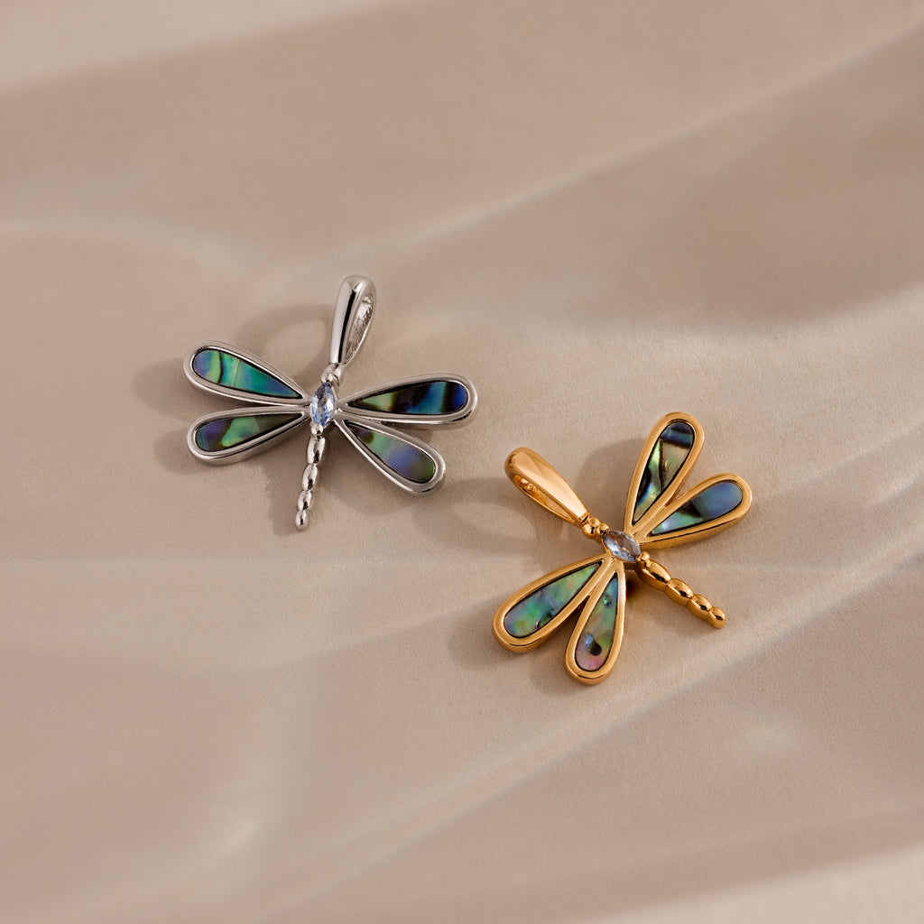 Two dragonfly-shaped pendants, one silver and one gold with iridescent wings, rest on a beige surface—capturing the nature-inspired beauty of the Abalone Dragonfly Charm.