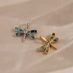 Two dragonfly-shaped pendants, one silver and one gold with iridescent wings, rest on a beige surface—capturing the nature-inspired beauty of the Abalone Dragonfly Charm.