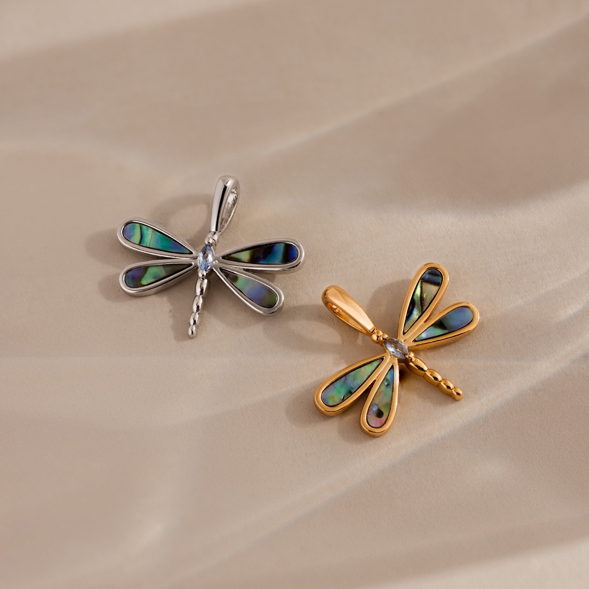 Two dragonfly-shaped pendants, one silver and one gold with iridescent wings, rest on a beige surface—capturing the nature-inspired beauty of the Abalone Dragonfly Charm.