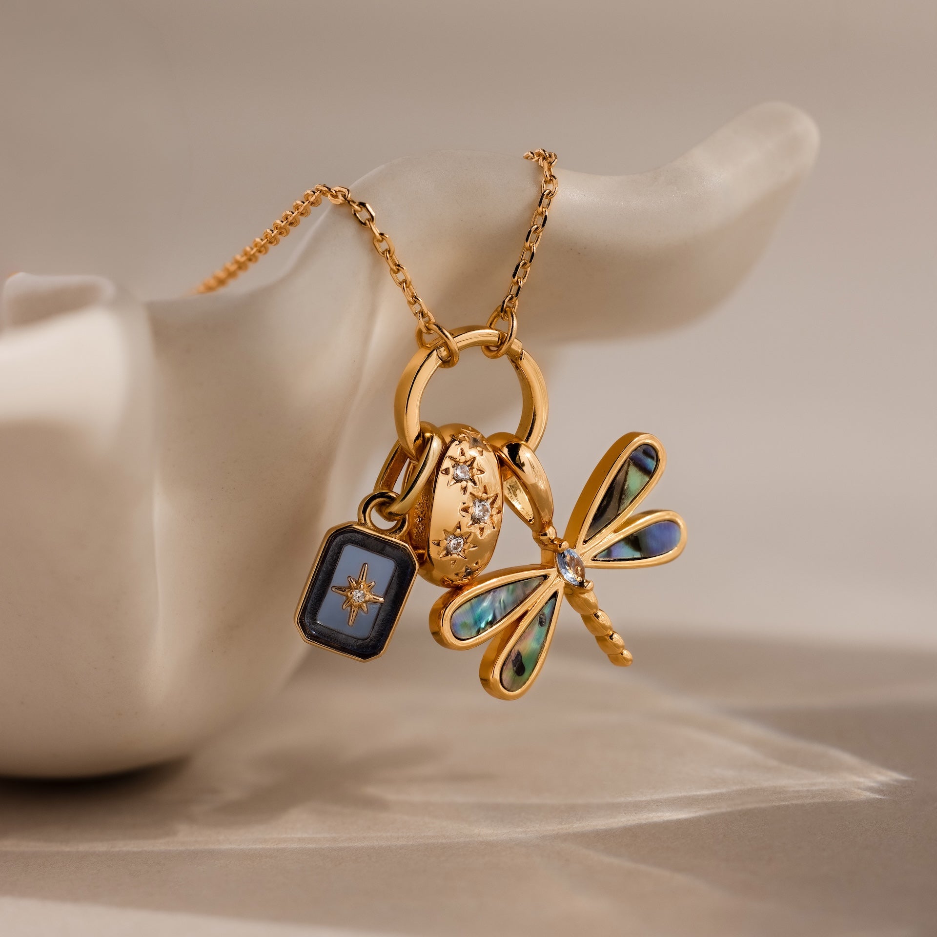 The Abalone Dragonfly Charm, a gold necklace featuring iridescent wing details, is displayed on a white sculpted ceramic hand.