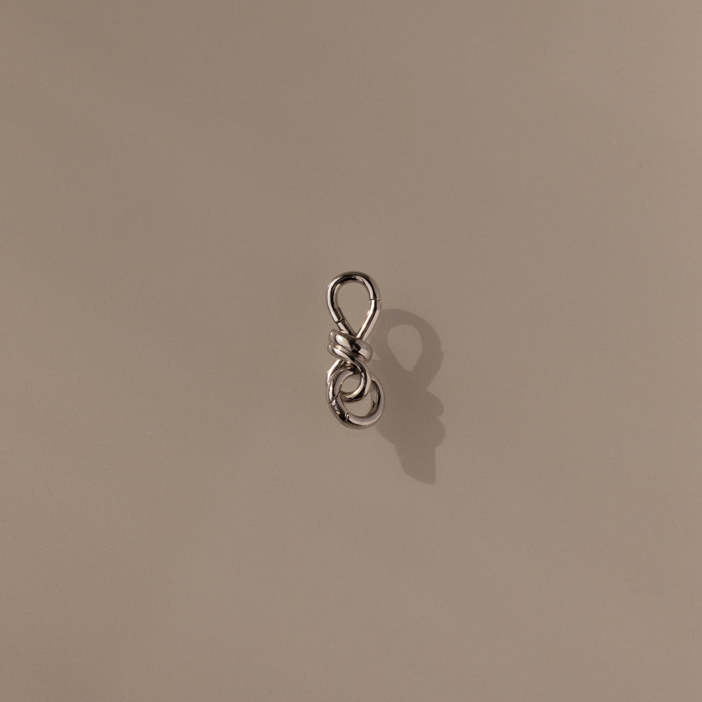 The Knot Charm Clasp in Silver, expertly photographed on a plain beige background, is a small metal clasp with a loop, perfect for jewelry making or crafts.