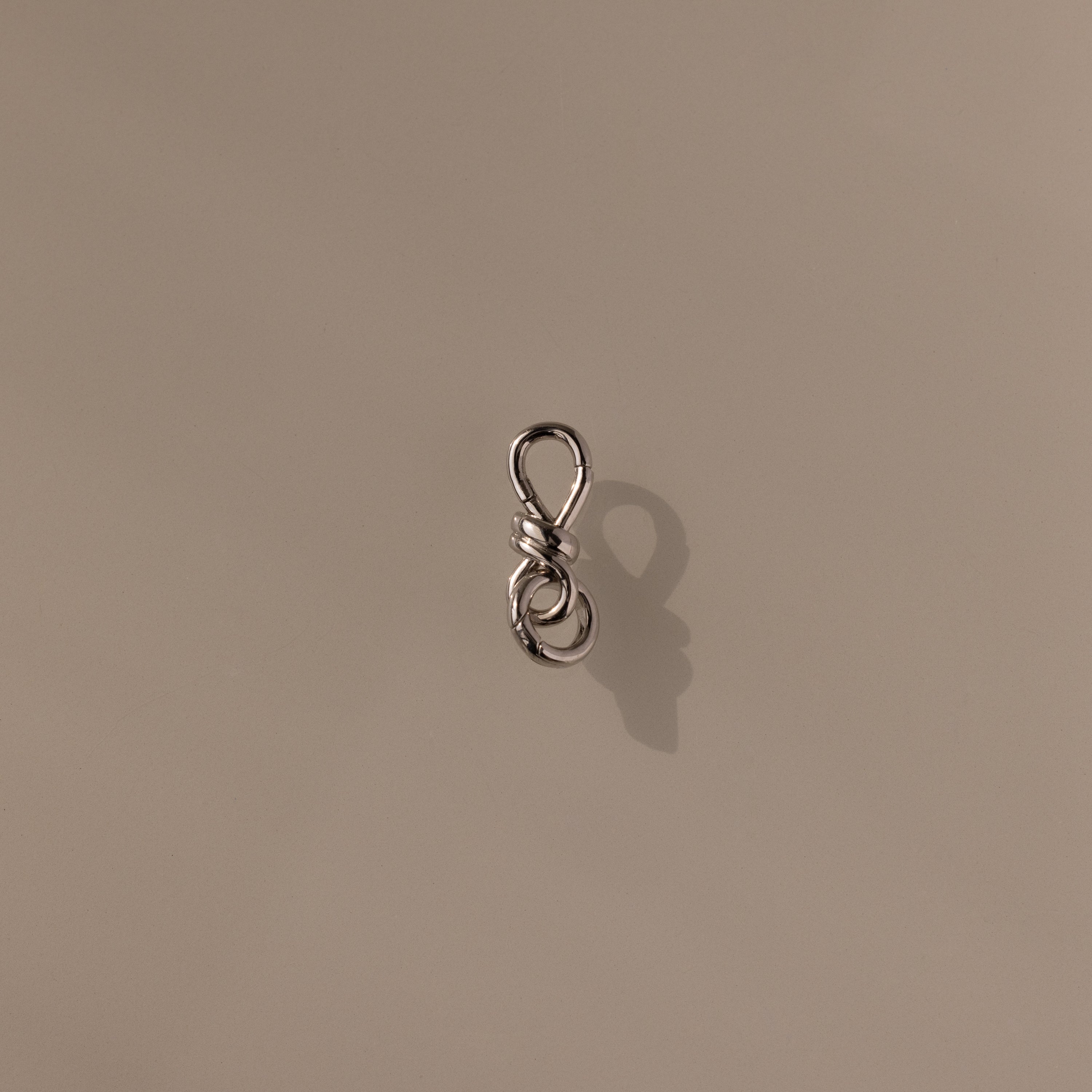 The Knot Charm Clasp in Silver, expertly photographed on a plain beige background, is a small metal clasp with a loop, perfect for jewelry making or crafts.