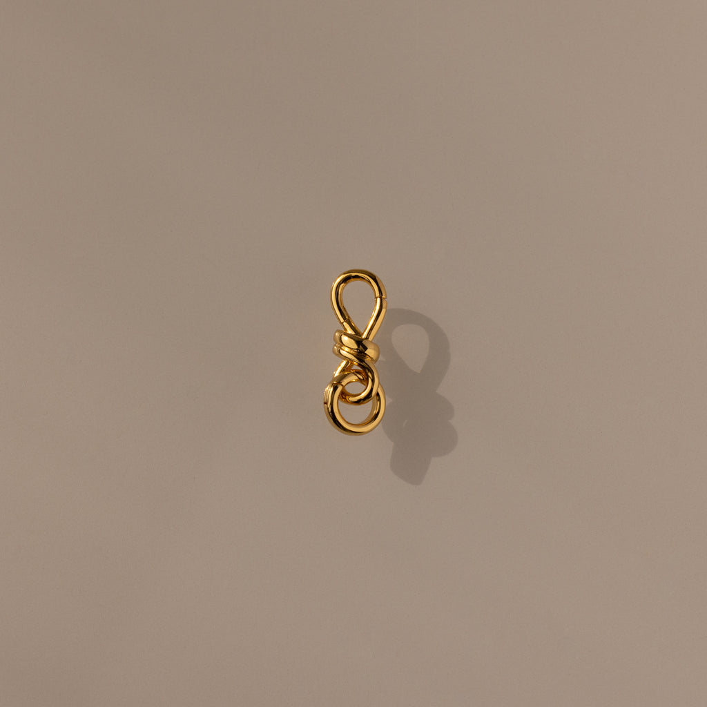 The Knot Charm Clasp in 18K Gold features a delicate gold wire shaped into a loop and knotted design, casting an elegant shadow on a plain beige surface—perfect for adding luxury and unique style to your jewelry collection.