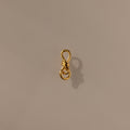 The Knot Charm Clasp in 18K Gold features a delicate gold wire shaped into a loop and knotted design, casting an elegant shadow on a plain beige surface—perfect for adding luxury and unique style to your jewelry collection.