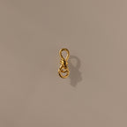 The Knot Charm Clasp in 18K Gold features a delicate gold wire shaped into a loop and knotted design, casting an elegant shadow on a plain beige surface—perfect for adding luxury and unique style to your jewelry collection.