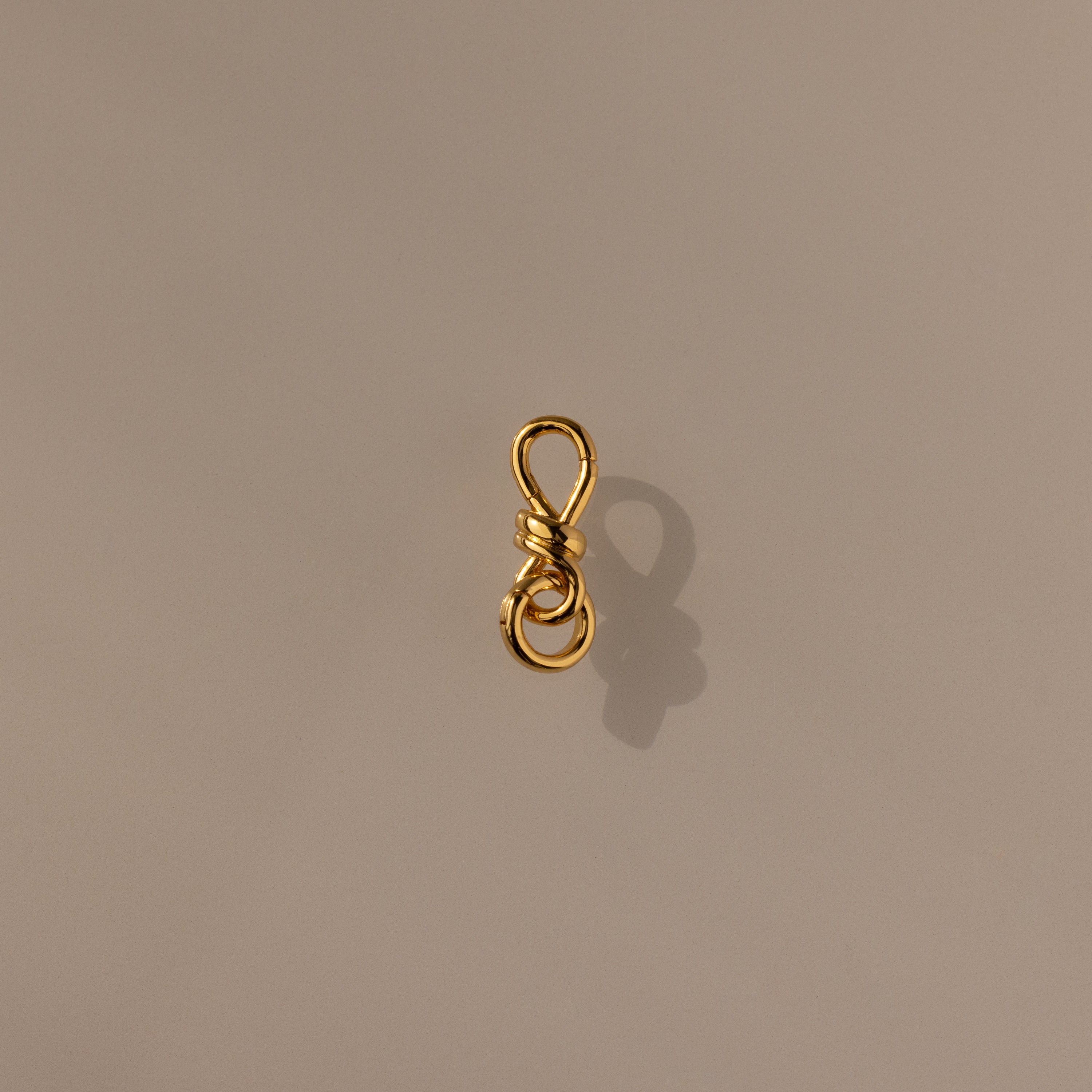 The Knot Charm Clasp in 18K Gold features a delicate gold wire shaped into a loop and knotted design, casting an elegant shadow on a plain beige surface—perfect for adding luxury and unique style to your jewelry collection.