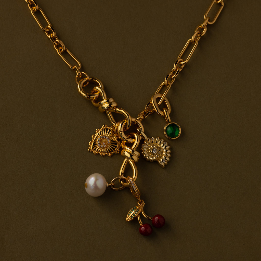 The Knot Charm Clasp in 18K Gold features whimsical flower, pearl, green gem, leaf, and cherry charms, beautifully displayed on an olive background.