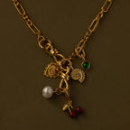 The Knot Charm Clasp in 18K Gold features whimsical flower, pearl, green gem, leaf, and cherry charms, beautifully displayed on an olive background.