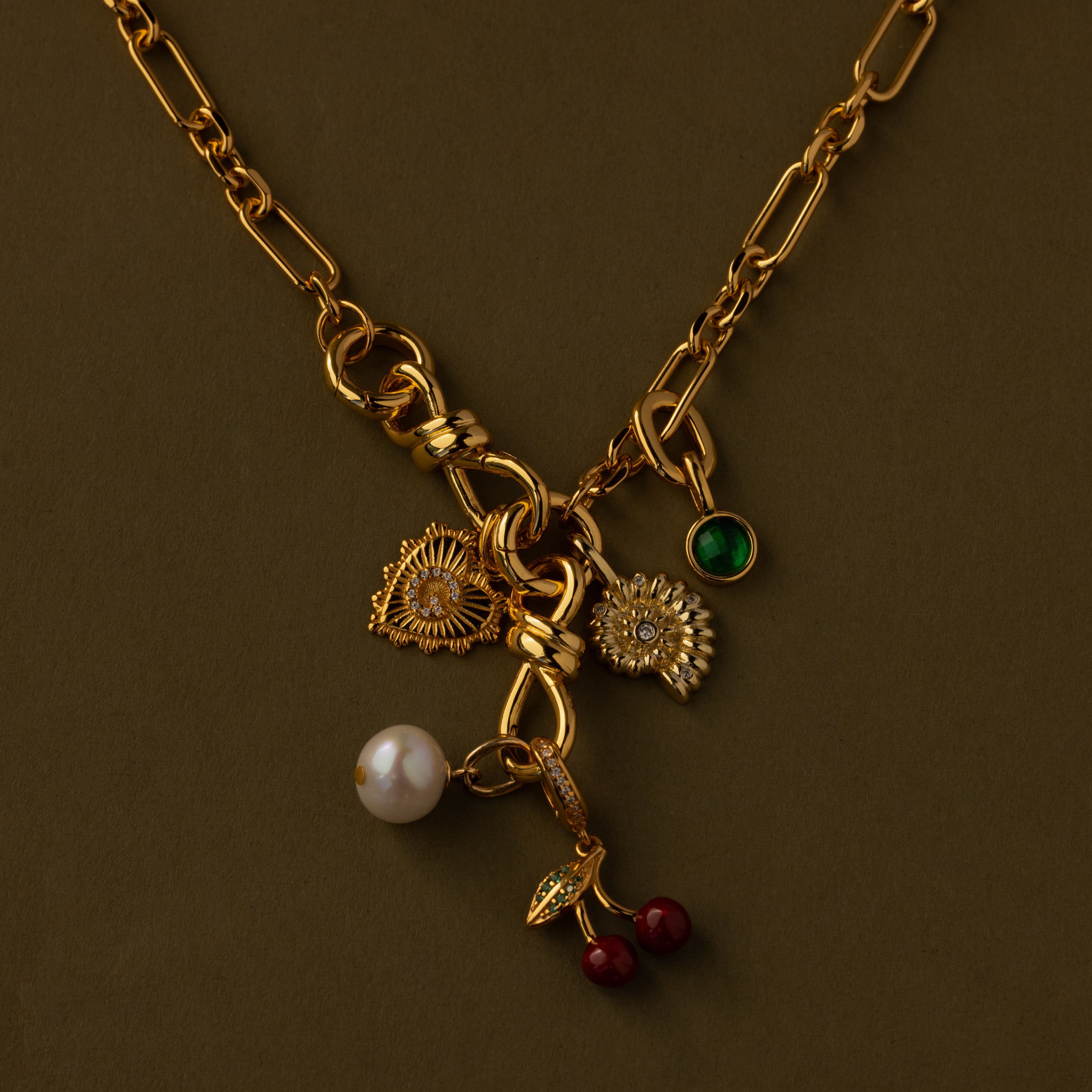 The Knot Charm Clasp in 18K Gold features whimsical flower, pearl, green gem, leaf, and cherry charms, beautifully displayed on an olive background.