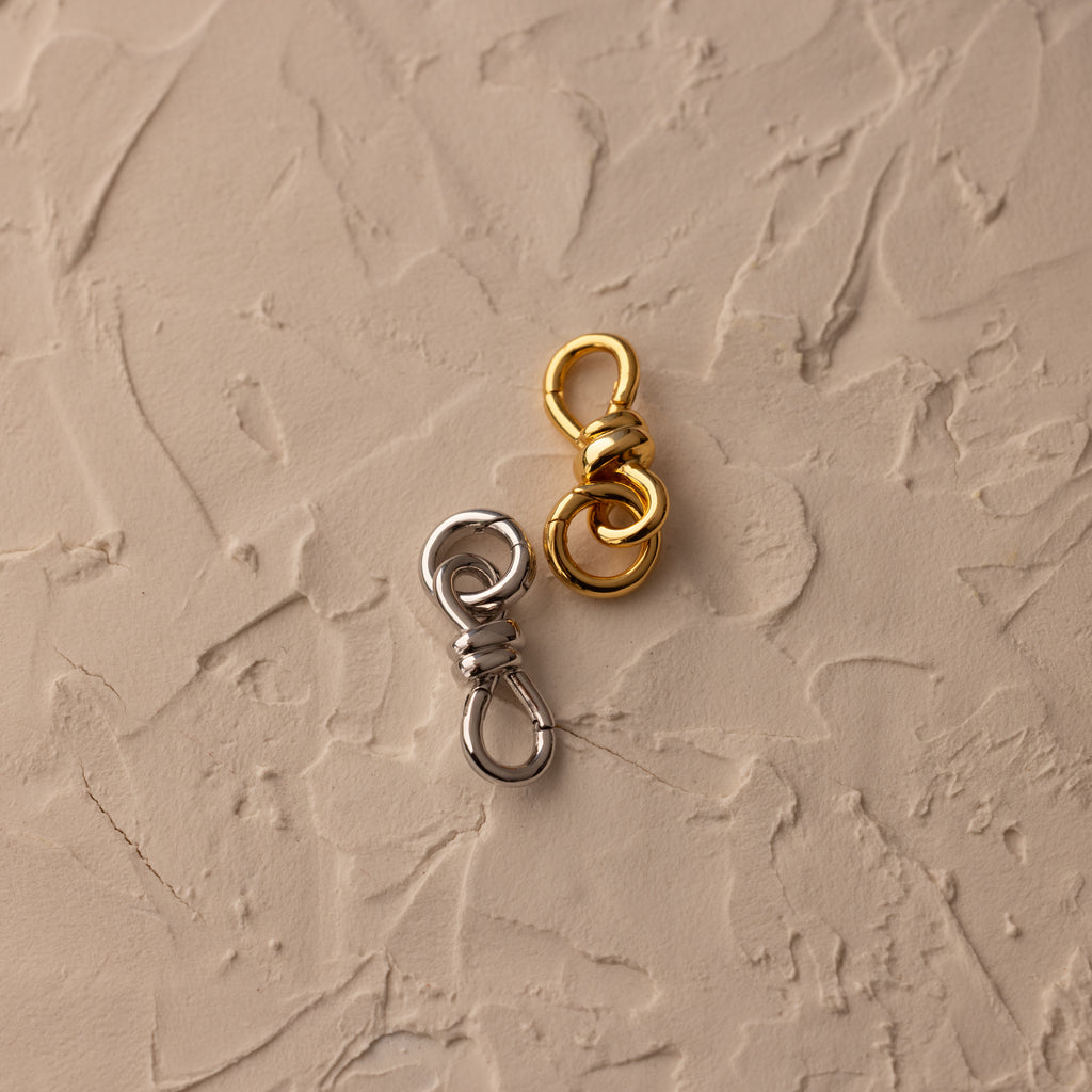 The Knot Charm Clasp in Silver is elegantly placed on a textured beige surface, showcasing its sleek metallic finish.