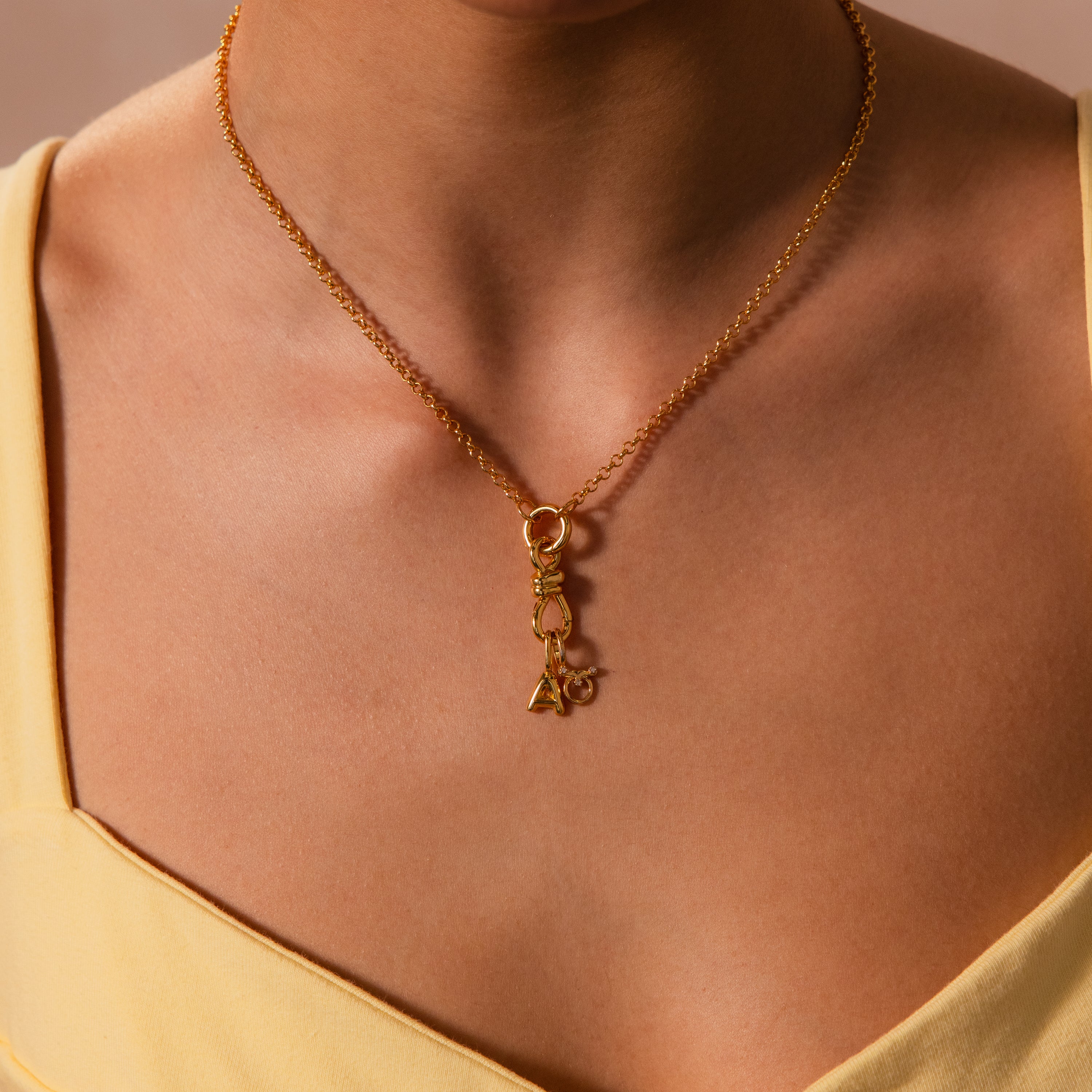 A person wears the Knot Charm Clasp in 18K Gold—a gold necklace featuring small geometric and letter charms—paired with a yellow top.