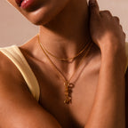 A woman wearing a cream top and layered gold necklaces, including the Knot Charm Clasp in Silver with a pendant, touches her neck.