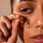 A woman holds a Knot Charm Clasp in 18K Gold close to her face, highlighting its elegant, intricate design and radiant gold finish.