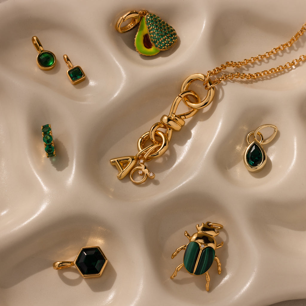 The Knot Charm Clasp, a gold and green jewelry piece, is displayed on a wavy cream surface—ideal for adding elegance to your accessory collection.