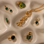 The Knot Charm Clasp, a gold and green jewelry piece, is displayed on a wavy cream surface—ideal for adding elegance to your accessory collection.