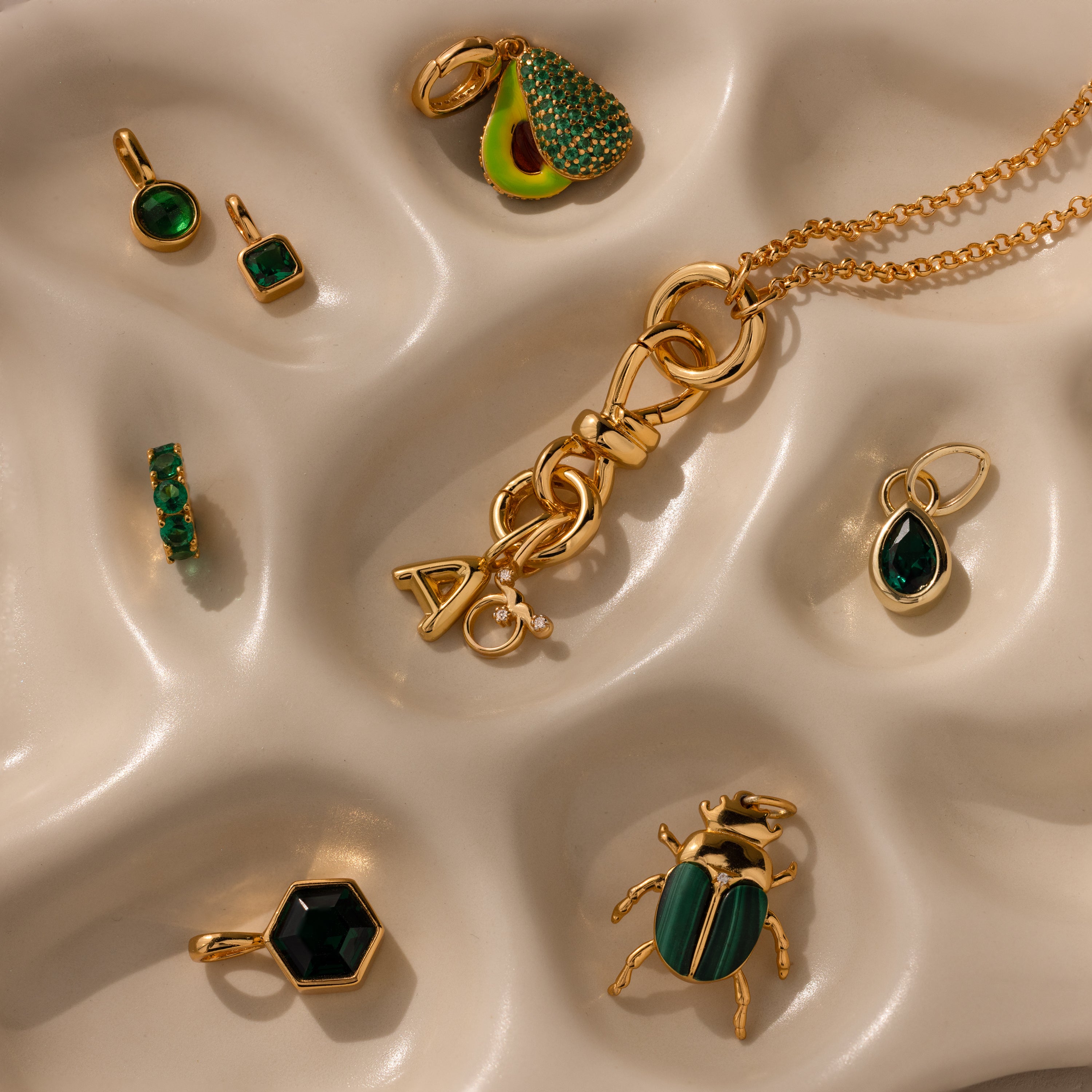 The Knot Charm Clasp, a gold and green jewelry piece, is displayed on a wavy cream surface—ideal for adding elegance to your accessory collection.
