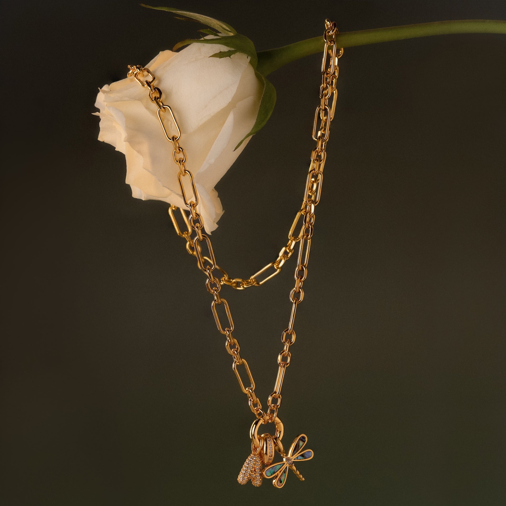 The Rolo Circle Figaro Necklace in 18K Gold features a butterfly and heart charm hanging from a white rose, set against a dark background, and showcases a classic link design for timeless elegance.