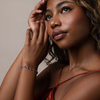 A woman in a red satin dress touches her face, gazing thoughtfully upward while wearing the Garnet Heart Tennis Bracelet in Sterling Silver—a stunning statement of elegance and allure.