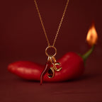 Gold necklace with a Chili Pepper Charm, red gem, and letter C, shown on a backdrop of red chili and flame.
