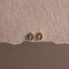 A pair of small, gold and silver stud earrings displayed on textured beige paper, these Rhombus Aquamarine Flatback Studs exude understated chic and modern glamour.