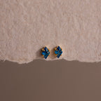 The Rhombus Blue Zircon Flatback Studs, featuring gold accents, offer understated chic with their small blue gemstones, elegantly displayed on a textured beige surface.