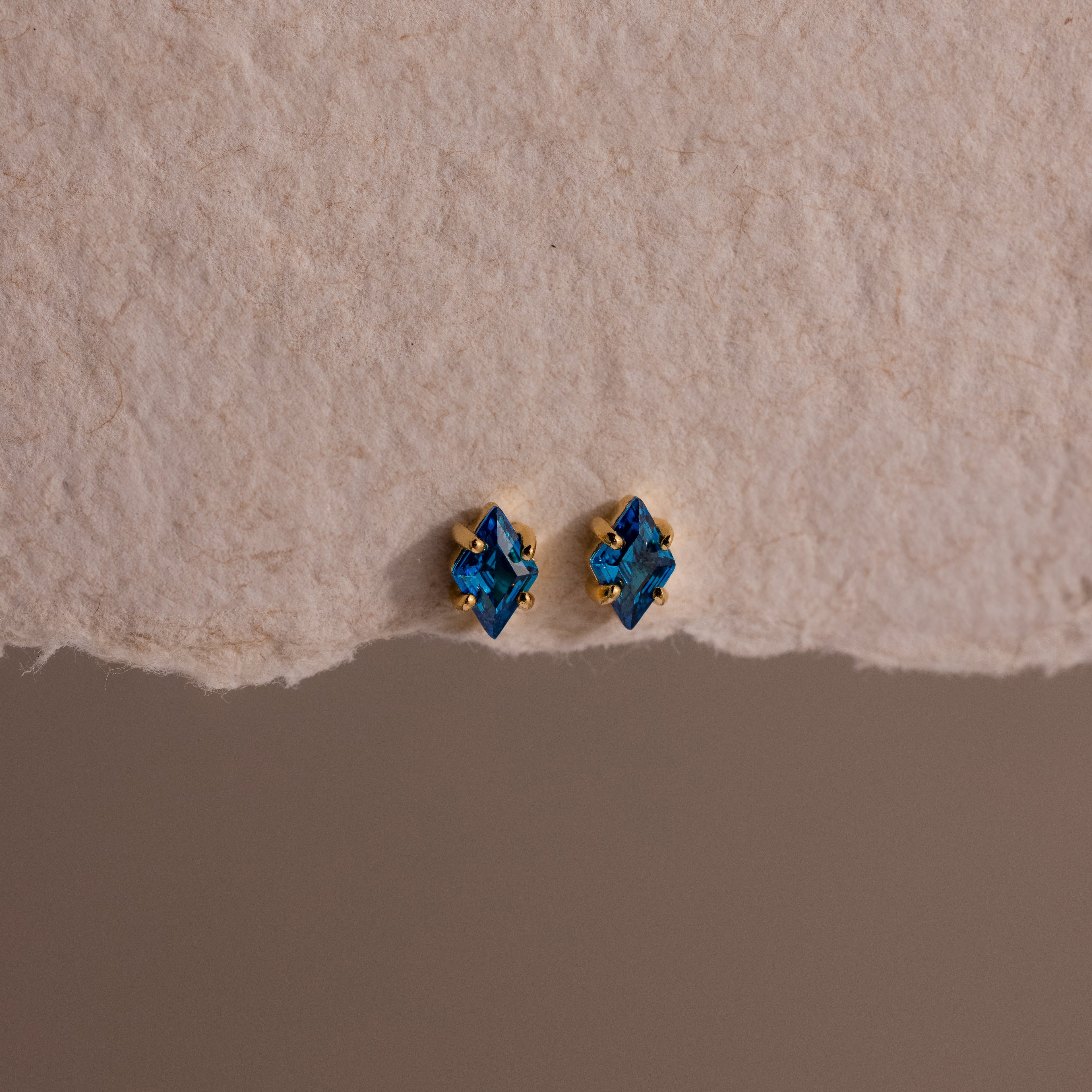 The Rhombus Blue Zircon Flatback Studs, featuring gold accents, offer understated chic with their small blue gemstones, elegantly displayed on a textured beige surface.