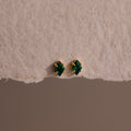 The Rhombus Emerald Flatback Studs are small gold earrings with green gemstone studs, embodying understated chic and presented on textured beige paper.
