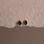 The Rhombus Emerald Flatback Studs are small gold earrings with green gemstone studs, embodying understated chic and presented on textured beige paper.