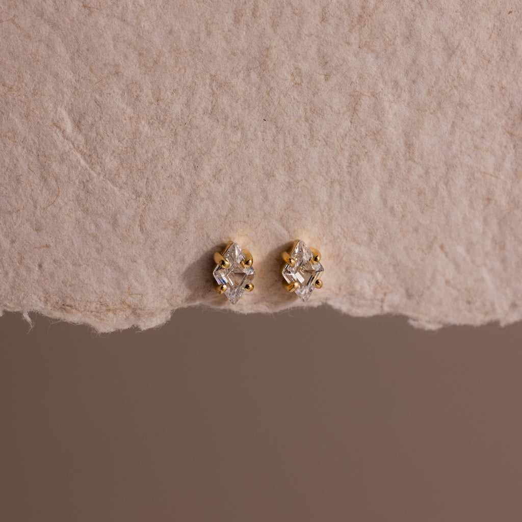 Rhombus Diamond Flatback Studs and other gold and diamond earrings are displayed on textured beige paper, creating an understated chic jewelry look.