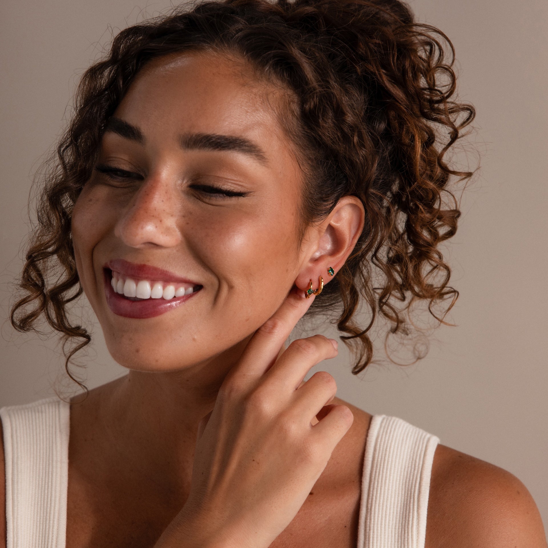 A smiling woman with curly hair touches her Rhombus Birthstone Flatback Studs, wearing a white sleeveless top against a neutral background.
