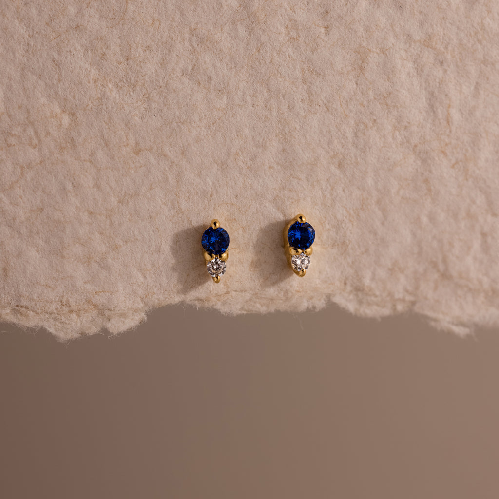 Tiny Duo Sapphire Flatback Studs—gold earrings with blue and clear stones, diamond accents, and shown on textured beige paper—perfect for your ear stack.