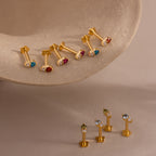 Six gold earrings with colorful gemstones on a beige surface, and four more upright below—perfect for showcasing vibrant ear stacks or highlighting Tiny Duo Blue Zircon Flatback Studs as bold birthstone accents.
