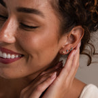 Smiling woman with curly hair touches her ear, showcasing chic Tiny Duo Pink Tourmaline Flatback Studs with delicate gold and gemstone accents in a stylish ear stack.