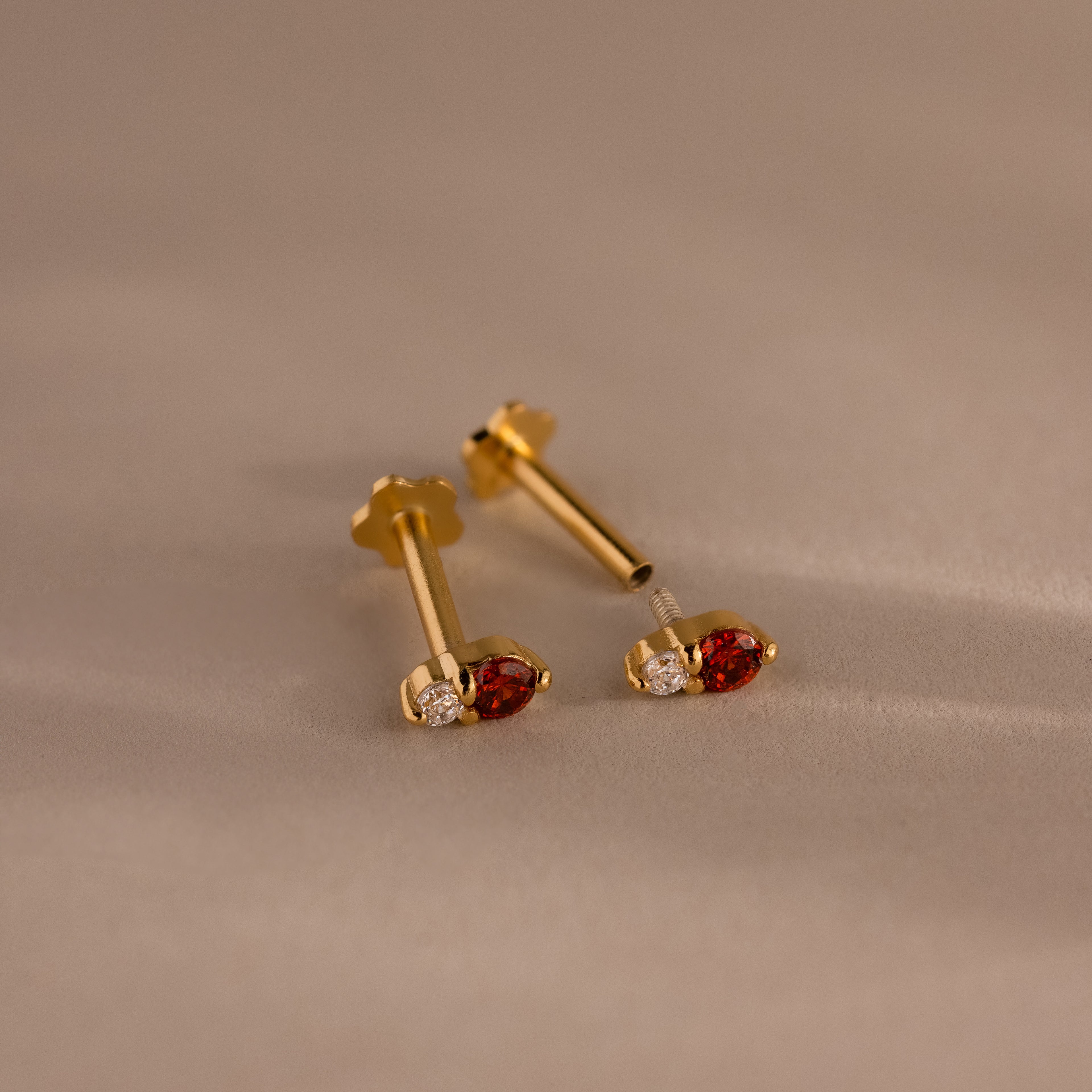 A pair of Tiny Duo Blue Zircon Flatback Studs with blue zircon stones and subtle diamond accents, ideal for an elegant ear stack, displayed on a beige surface.