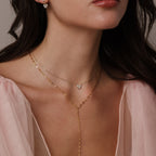 A woman wears the Mirror Chain Lariat Necklace in 18K Gold, its reflective finish layered over a light pink sheer top, highlighting her neck and lower face.