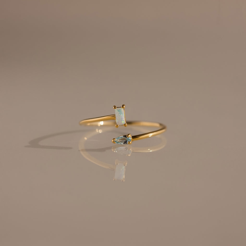 The Toi et Moi Wrap Ring in 18K Gold features two small opal stones and is displayed on a neutral beige surface.