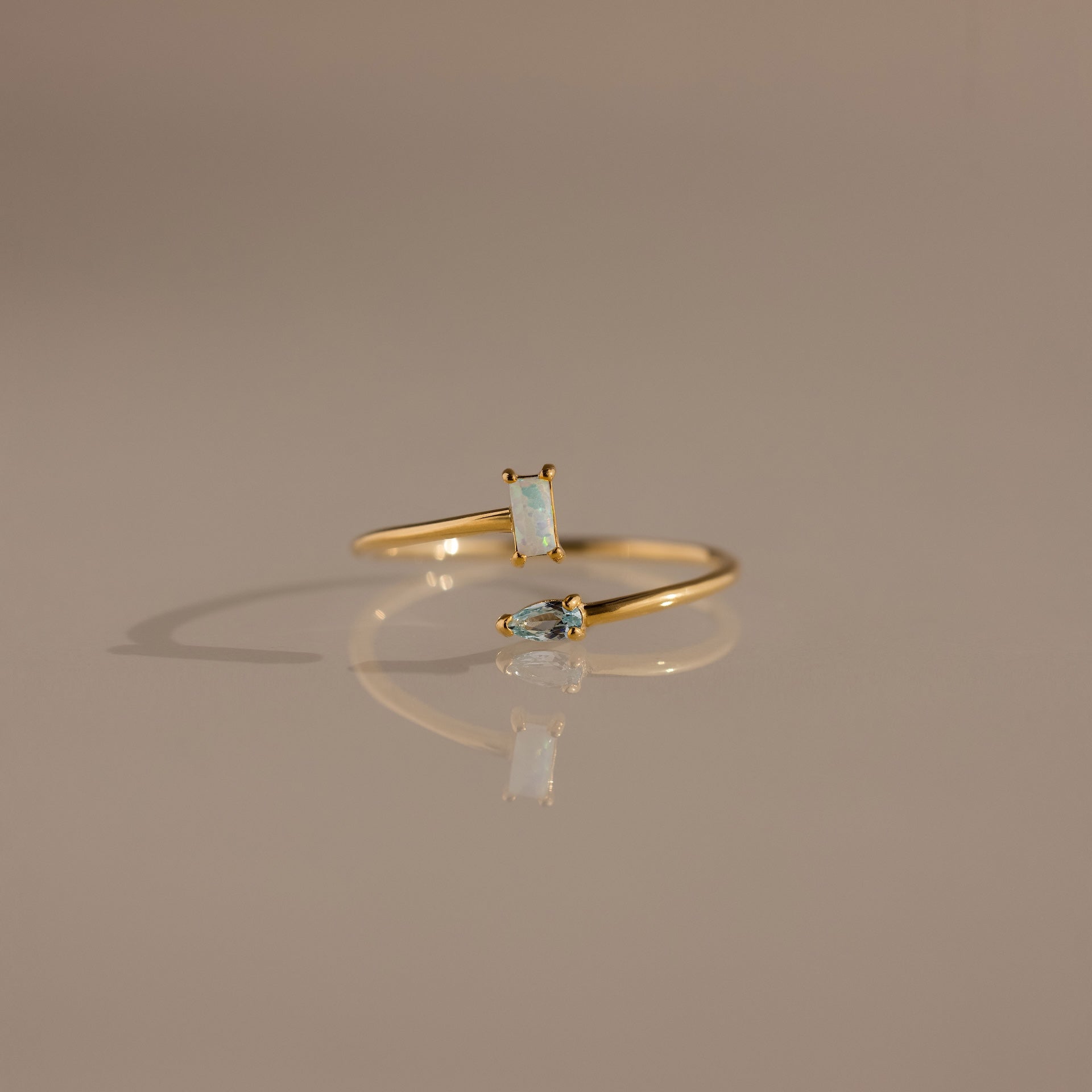 The Toi et Moi Wrap Ring in 18K Gold features two small opal stones and is displayed on a neutral beige surface.
