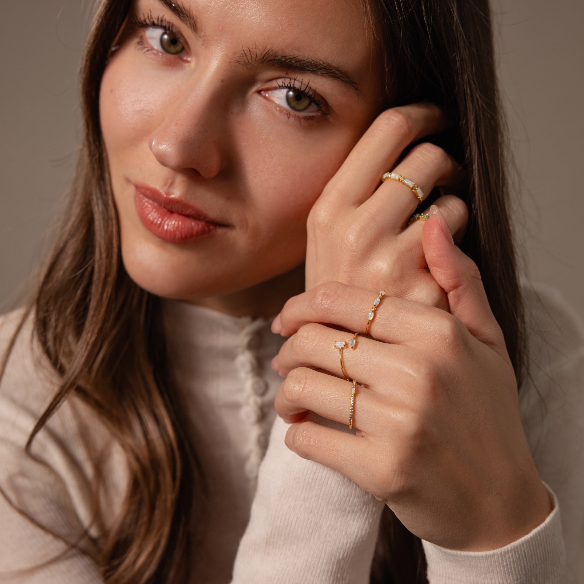 Woman with long brown hair wears a cream top and several gold rings, including the elegant Toi et Moi Wrap Ring in 18K Gold, as she holds her hands near her face.