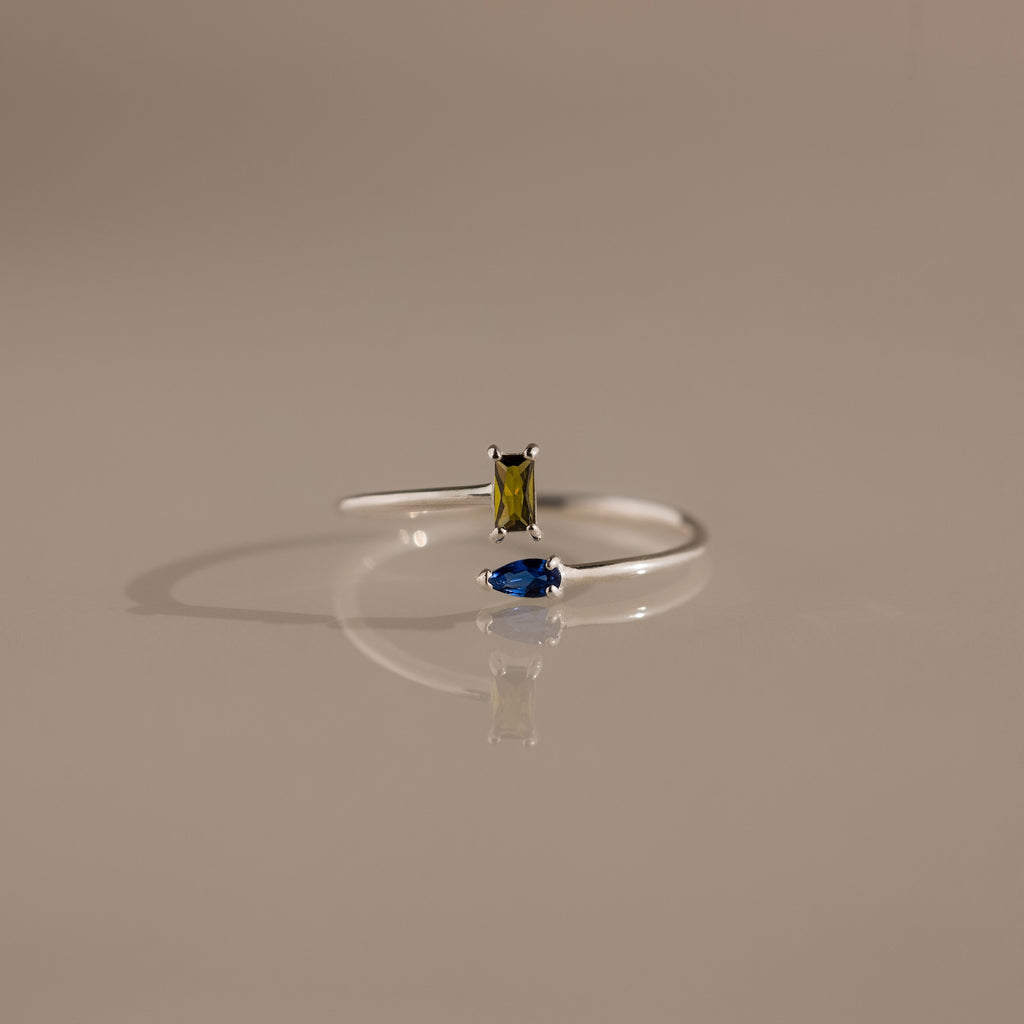 The Toi et Moi Wrap Ring features a rectangular yellow gemstone and a marquise blue gemstone set in silver, displayed on a neutral background—a timeless romantic jewelry piece.