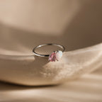 The Duo Pear Toi Et Moi Ring in Silver, featuring pink and white opal pear-shaped stones, is displayed on a smooth beige surface under soft lighting.