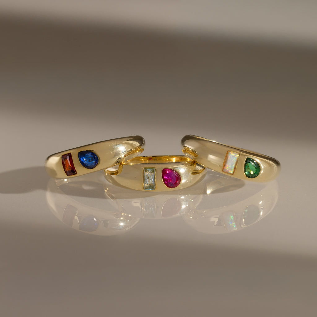 The Toi et Moi Dome Ring in 18K Gold, set with colorful blue, red, green, and iridescent gemstones, sparkles elegantly on a reflective surface.