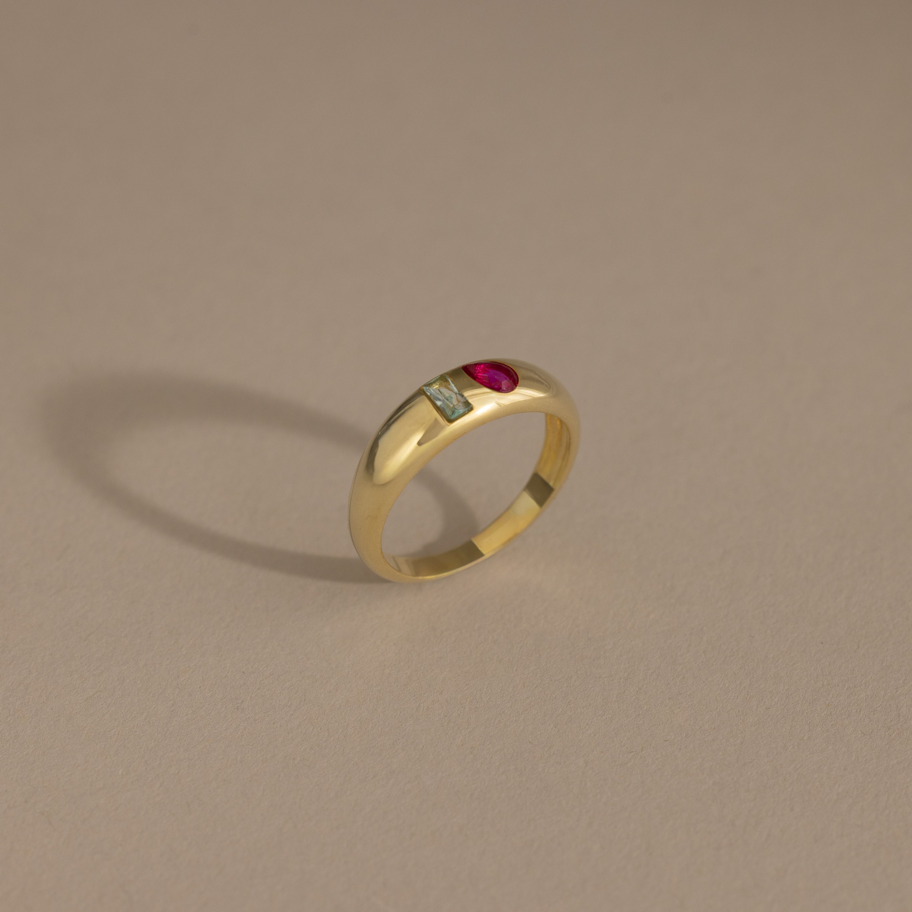 The Toi et Moi Dome Ring in 18K Gold features a rectangular green stone and a round red stone set side by side, displayed on a beige background.