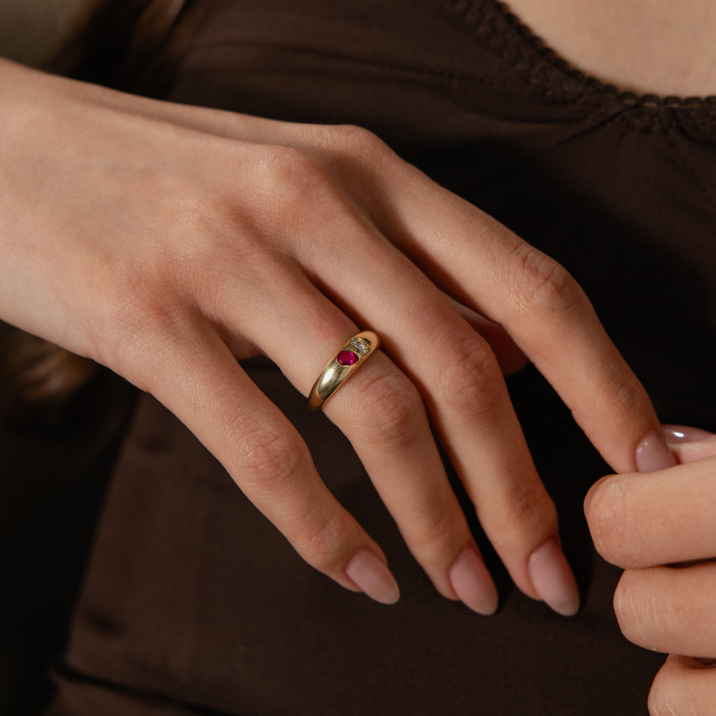 A hand with manicured nails wears the Toi et Moi Dome Ring, a gold birthstone ring with a red gemstone, resting on a brown top.