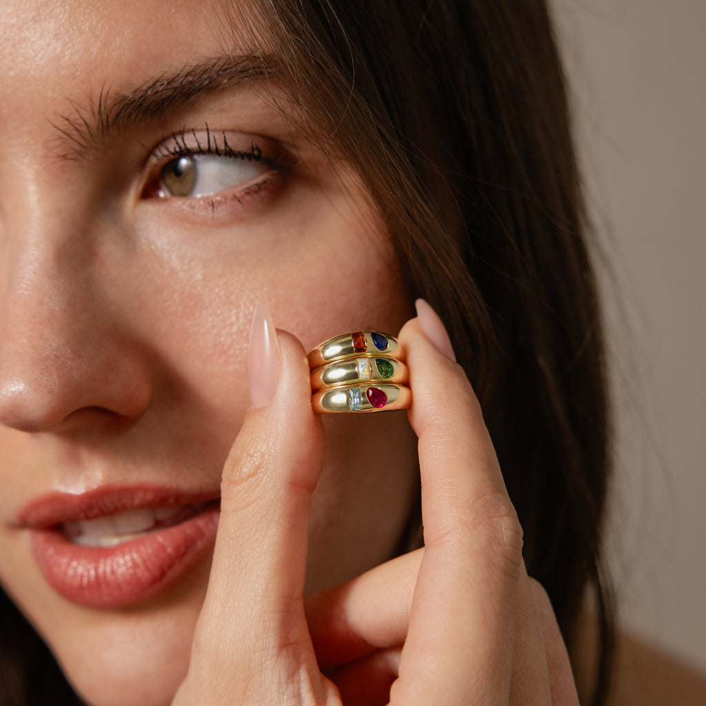 A woman holds up the Toi et Moi Dome Ring in 18K Gold with colorful gemstones near her face, highlighting its playful and trendy design to the camera.