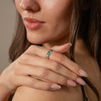 A woman with manicured nails touches her shoulder, highlighting the elegance of the Toi et Moi Dome Ring featuring vibrant blue and green stones—a modern take on classic birthstone jewelry.