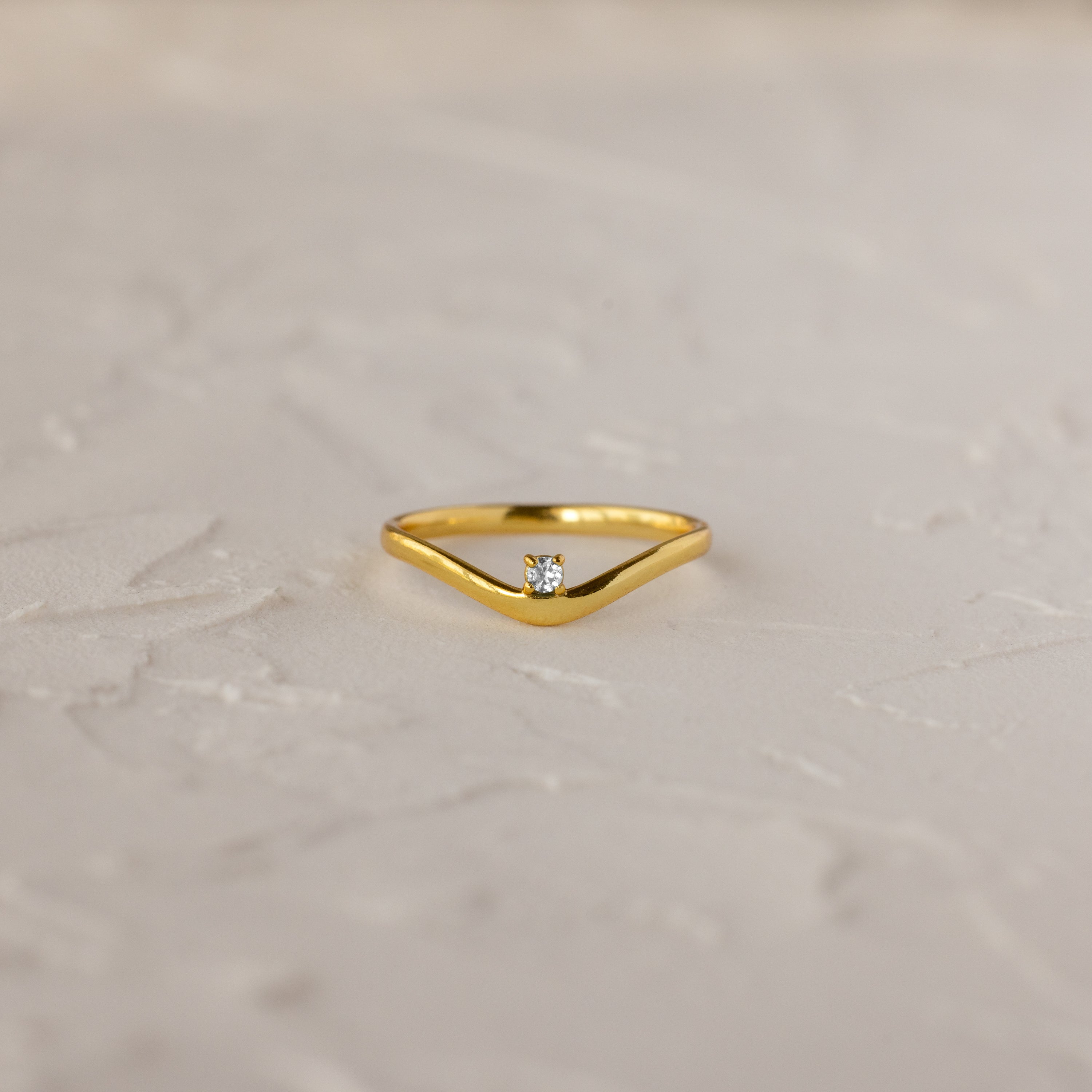 The Diamond Drop Curve Ring features a V-shaped gold band with a small central diamond, displayed on a textured light surface.