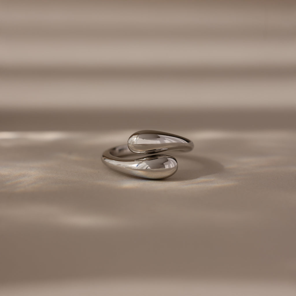 The Duo Dome Ring in White Gold, featuring an abstract sculptural design and adjustable band, rests on a beige surface under soft, diffused light—an elegant statement piece.