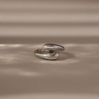 The Duo Dome Ring in White Gold, featuring an abstract sculptural design and adjustable band, rests on a beige surface under soft, diffused light—an elegant statement piece.
