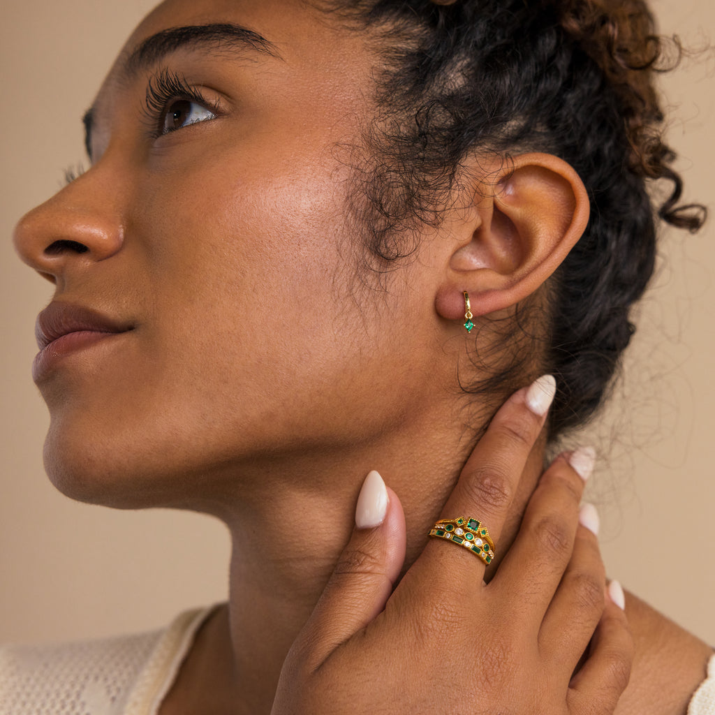 A woman with curly hair wears Emerald Drop Huggies and gold rings with green stones, touching her face gently to showcase personalized jewelry.