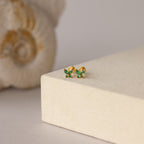 The Butterfly Emerald Flatback Studs—gold earrings with small green gemstones—add a personalized jewelry touch, shown on a beige platform beside a shell.